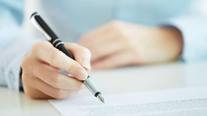 A person in a blue sweater signing a document on a clipboard with a silver pen